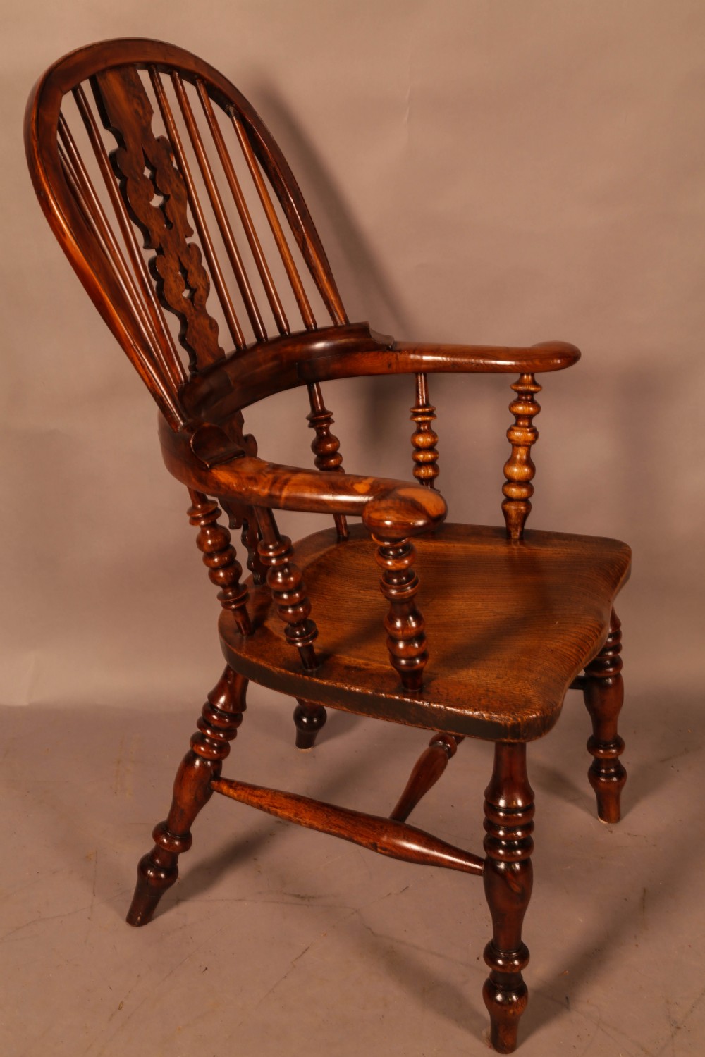 An antique brown yew wood Windsor armchair featuring a hoop back, a central pierced splat, turned spindles, broad armrests, and turned legs.