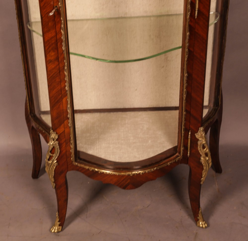 An upright curved glass display cabinet veneered in rich brown kingwood with intricate cast ormolu mounts and glass shelving. View IV
