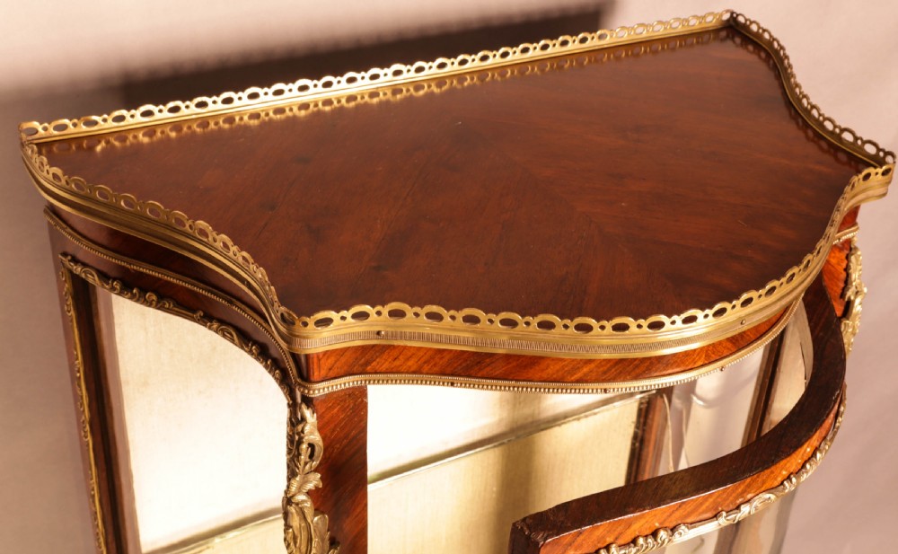 An upright curved glass display cabinet veneered in rich brown kingwood with intricate cast ormolu mounts and glass shelving. View VI