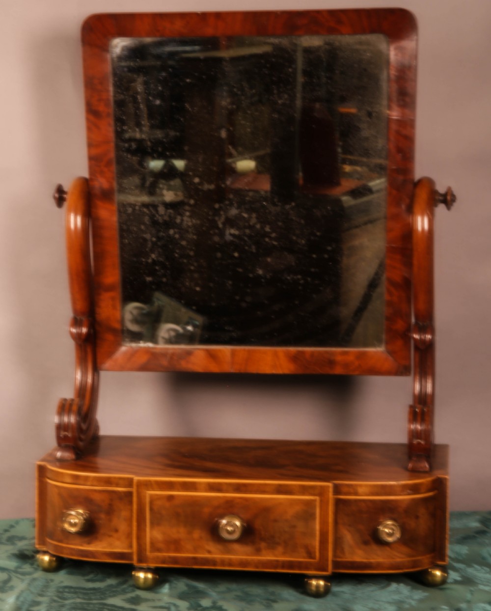 An early Victorian flame mahogany dressing mirror with a rectangular swivelling glass frame on scroll supports above a plinth drawer base with brass feet.
