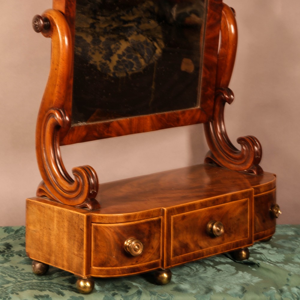 An early Victorian flame mahogany dressing mirror with a rectangular swivelling glass frame on scroll supports above a plinth drawer base with brass feet. View II