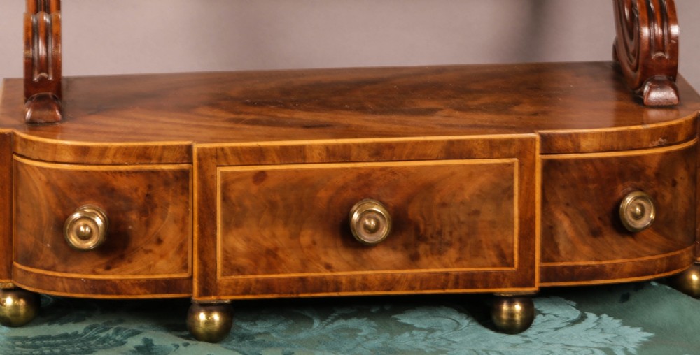 An early Victorian flame mahogany dressing mirror with a rectangular swivelling glass frame on scroll supports above a plinth drawer base with brass feet. View III