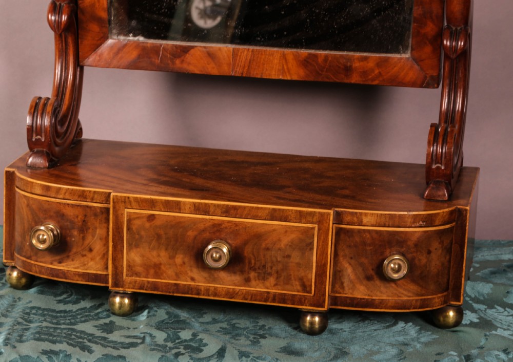 An early Victorian flame mahogany dressing mirror with a rectangular swivelling glass frame on scroll supports above a plinth drawer base with brass feet. View IV