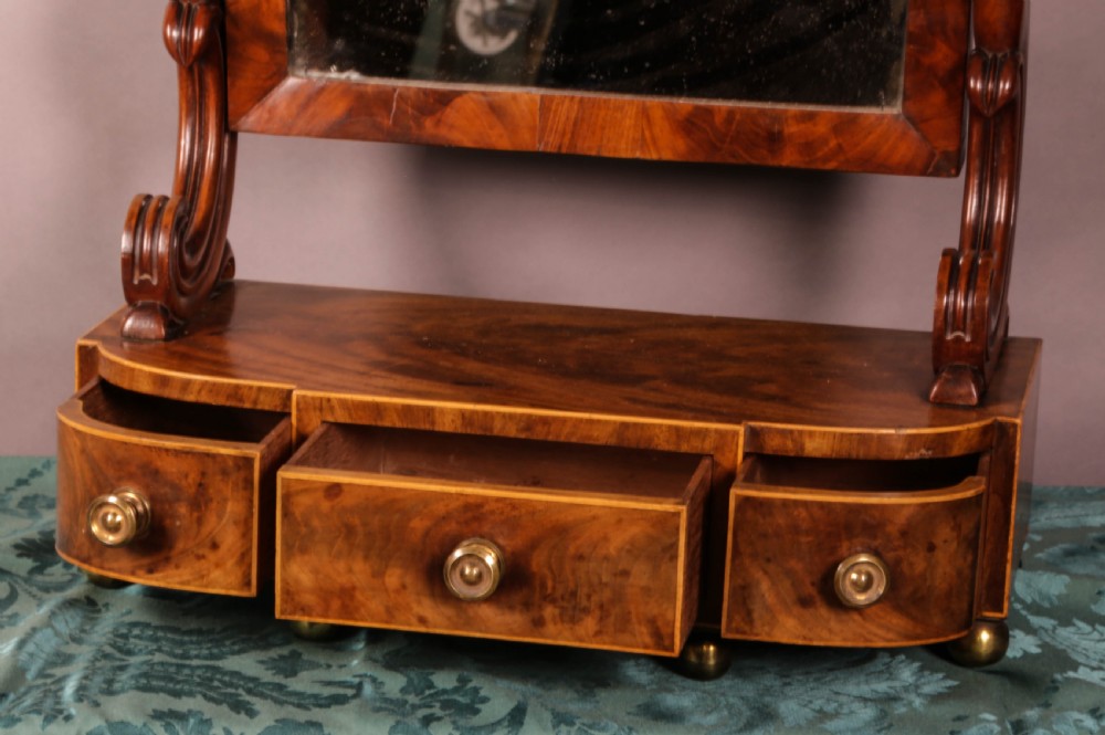An early Victorian flame mahogany dressing mirror with a rectangular swivelling glass frame on scroll supports above a plinth drawer base with brass feet. View V