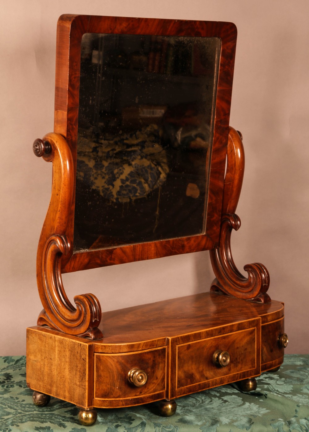 An early Victorian flame mahogany dressing mirror with a rectangular swivelling glass frame on scroll supports above a plinth drawer base with brass feet. View VI