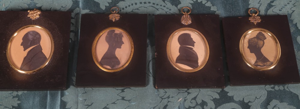 Four framed antique silhouette profile portraits in rectangular papier-mache frames with oval brass bezels and decorative metal hangers.
