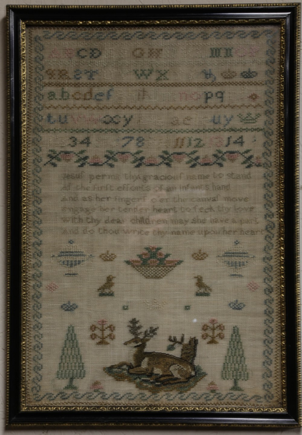 A framed 19th-century needlework sampler on linen featuring embroidered alphabets, numbers, a central verse, floral borders, and a reclining stag motif.