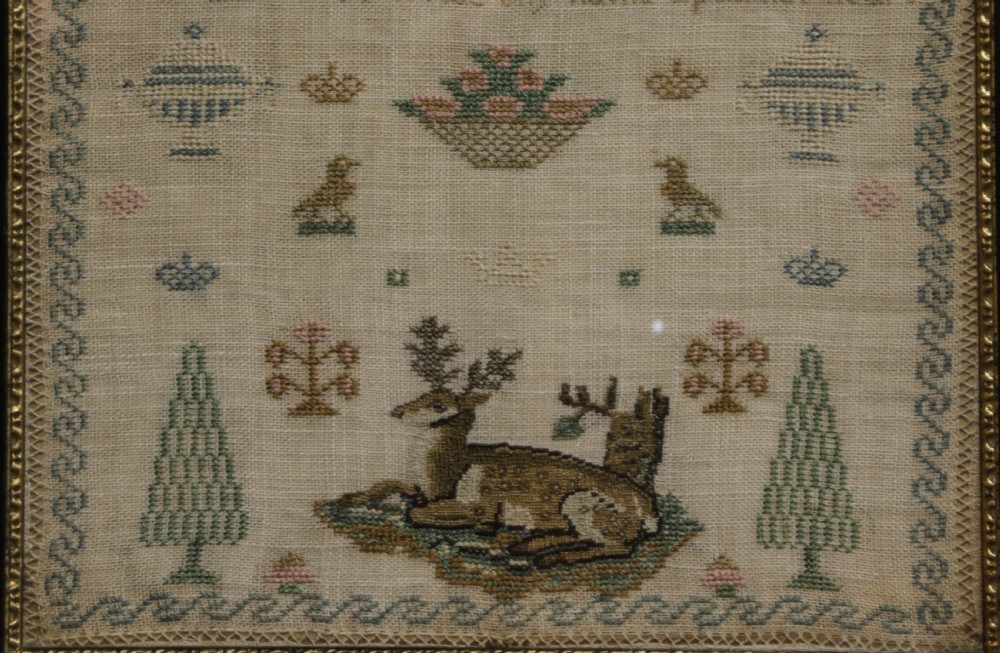A framed 19th-century needlework sampler on linen featuring embroidered alphabets, numbers, a central verse, floral borders, and a reclining stag motif. View IV