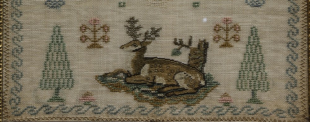 A framed 19th-century needlework sampler on linen featuring embroidered alphabets, numbers, a central verse, floral borders, and a reclining stag motif. View V