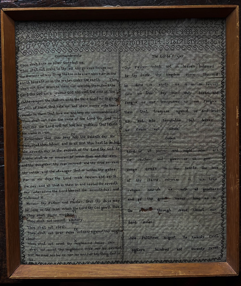 An 1827 Georgian embroidered needlework sampler featuring the Ten Commandments, the Lords Prayer, and an alphabet border set in a wooden frame.