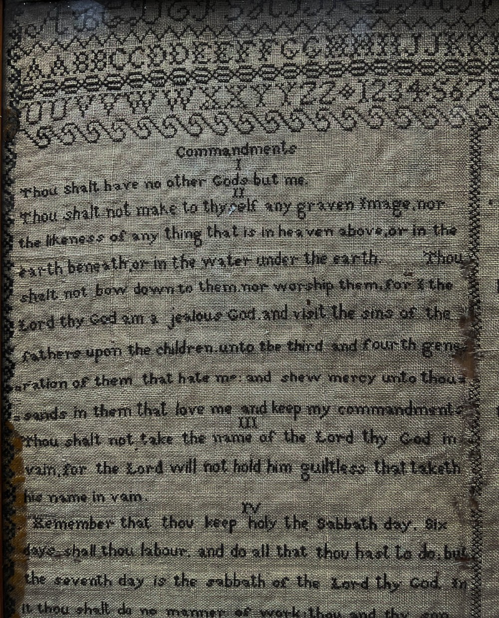An 1827 Georgian embroidered needlework sampler featuring the Ten Commandments, the Lords Prayer, and an alphabet border set in a wooden frame. View II