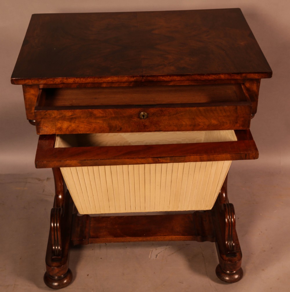 An antique William IV mahogany sewing table featuring a rectangular top, a single frieze drawer with a brass keyhole, a pleated fabric work bag suspended below, and scroll-carved end supports resting on bun feet. View II