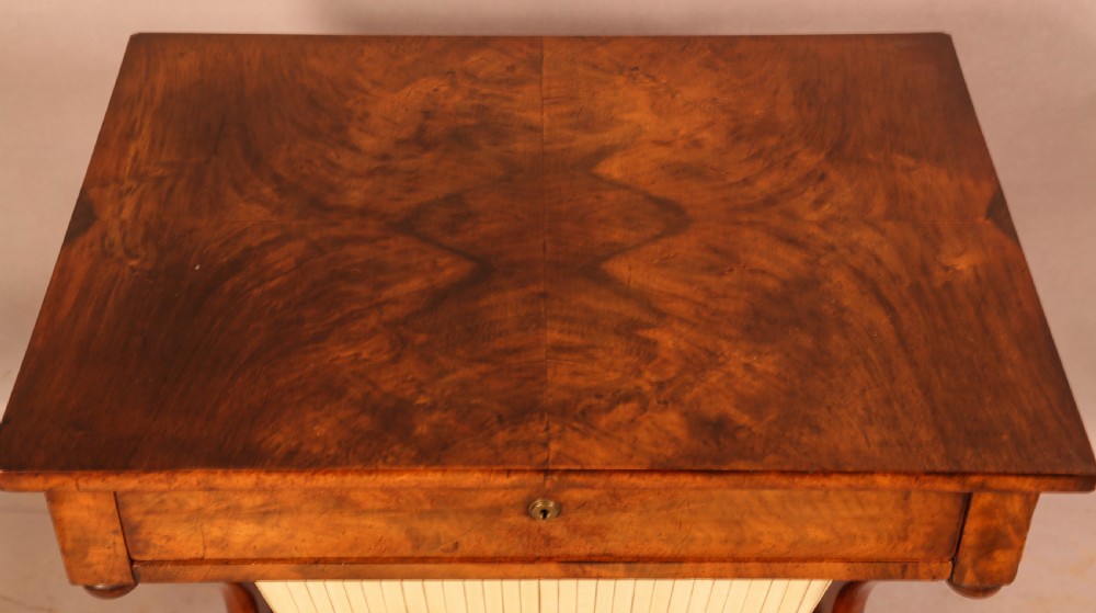 An antique William IV mahogany sewing table featuring a rectangular top, a single frieze drawer with a brass keyhole, a pleated fabric work bag suspended below, and scroll-carved end supports resting on bun feet. View III