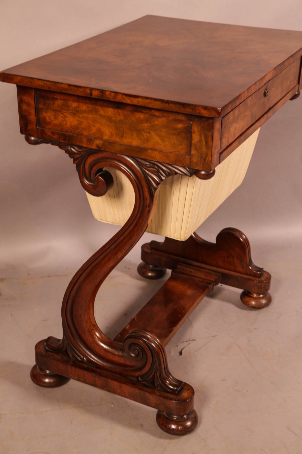 An antique William IV mahogany sewing table featuring a rectangular top, a single frieze drawer with a brass keyhole, a pleated fabric work bag suspended below, and scroll-carved end supports resting on bun feet. View IV