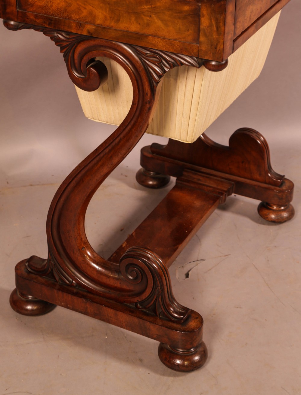 An antique William IV mahogany sewing table featuring a rectangular top, a single frieze drawer with a brass keyhole, a pleated fabric work bag suspended below, and scroll-carved end supports resting on bun feet. View V