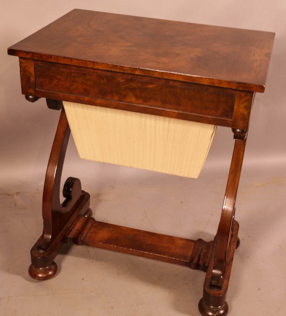 An antique William IV mahogany sewing table featuring a rectangular top, a single frieze drawer with a brass keyhole, a pleated fabric work bag suspended below, and scroll-carved end supports resting on bun feet. View VII