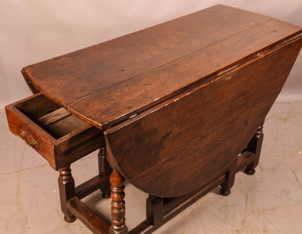 A seventeenth-century oak gateleg dining table featuring two drop leaves, turned legs, perimeter stretchers, and an open end drawer.