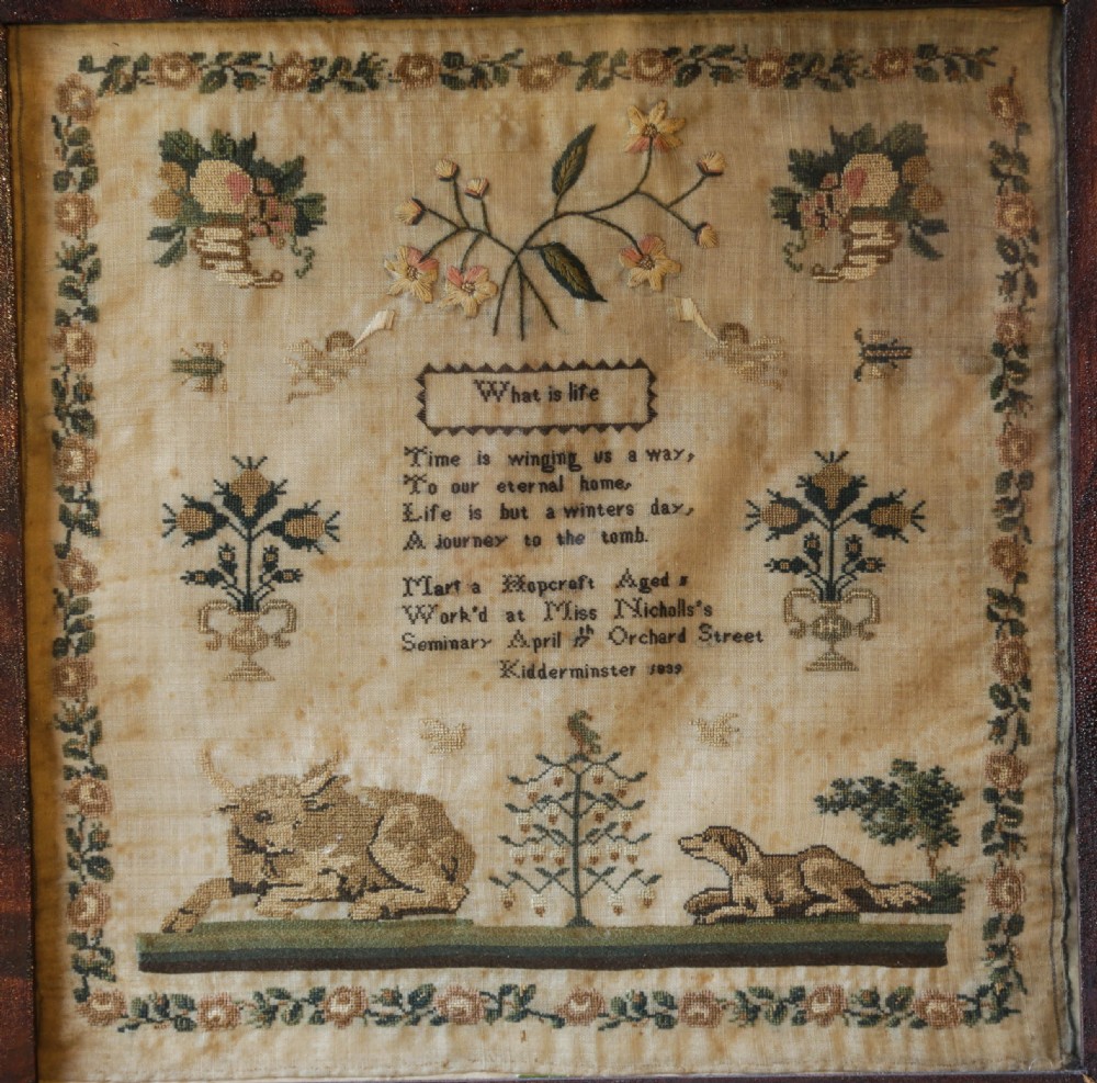 A framed antique needlework sampler featuring embroidered verses, flora, fauna, and figures on an aged linen ground.