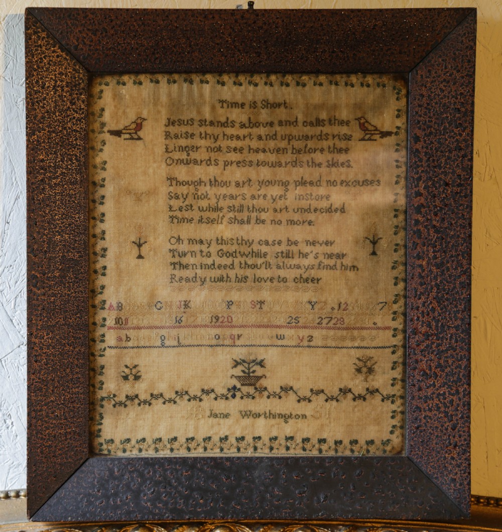 A framed antique needlework sampler on linen featuring stitched alphabets, numbers, verses, and motifs, enclosed in a dark textured wooden frame.