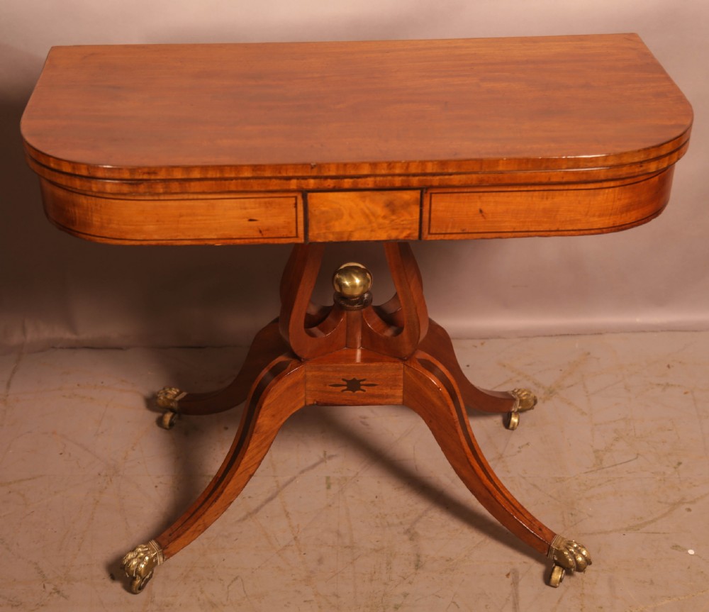 An antique Regency mahogany fold-over games table with a curved frieze, central pedestal support, and splayed legs with brass paw feet.