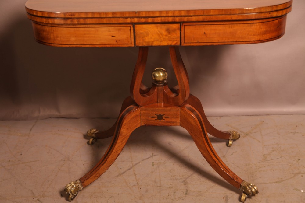 An antique Regency mahogany fold-over games table with a curved frieze, central pedestal support, and splayed legs with brass paw feet. View II