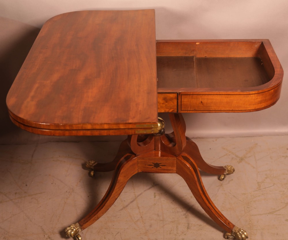 An antique Regency mahogany fold-over games table with a curved frieze, central pedestal support, and splayed legs with brass paw feet. View X