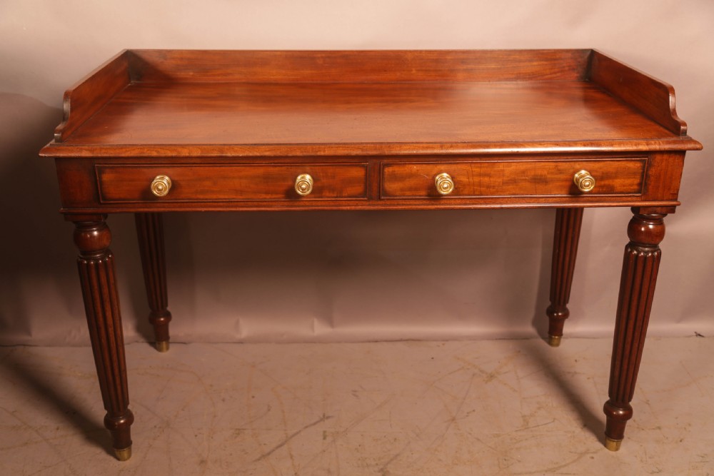 A rectangular mahogany library table featuring a raised back gallery, four drawers with brass ring pulls, and fluted tapering legs.