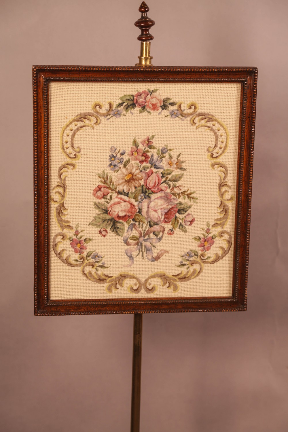 A nineteenth-century dark wood pole screen featuring an embroidered floral tapestry panel mounted on a brass rod with a heavily carved tripod base. View II
