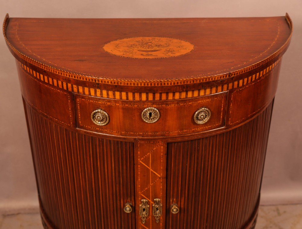 An eighteenth-century mahogany demi-lune cabinet featuring intricate geometric inlay, tambour doors, and brass hardware. View V