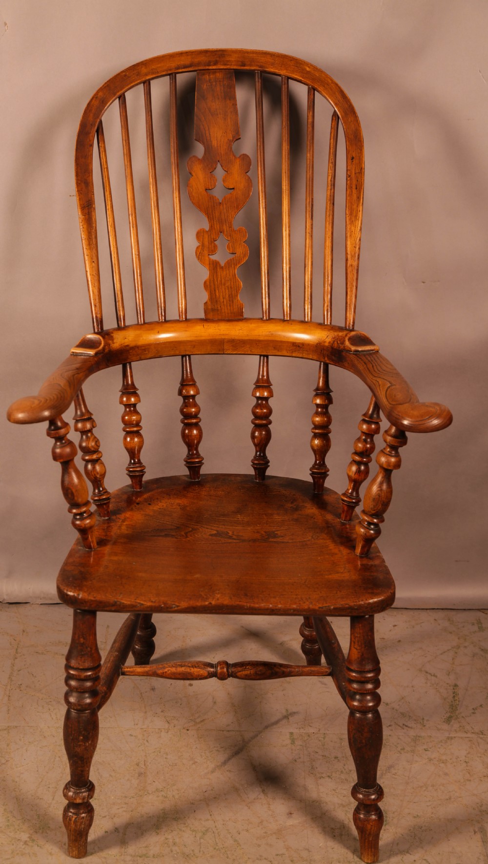 A Victorian broad arm Windsor armchair made from warm brown ash and elm timber, featuring a pierced central splat, turned spindles, a contoured saddle seat, and turned legs with stretchers.