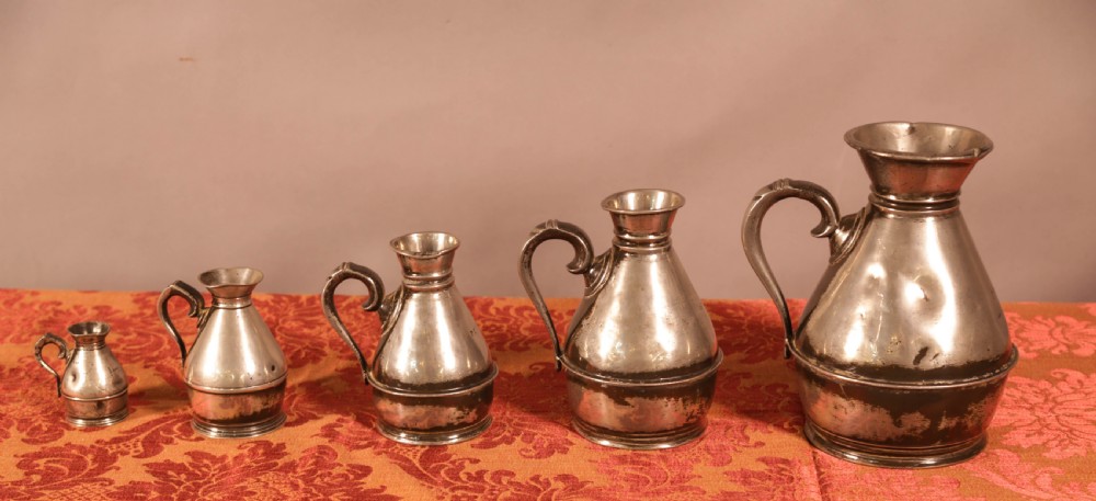 A graduated set of five antique pewter harvest jugs lined up on a patterned red surface.