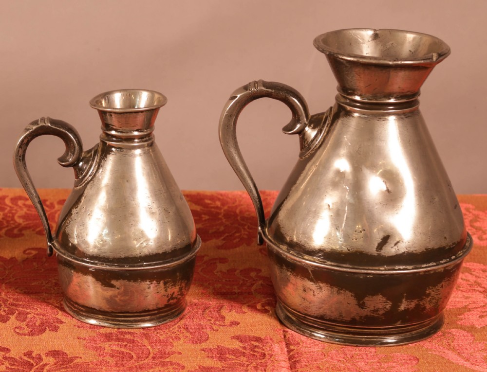 A graduated set of five antique pewter harvest jugs lined up on a patterned red surface. View II
