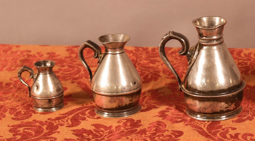 A graduated set of five antique pewter harvest jugs lined up on a patterned red surface. View III