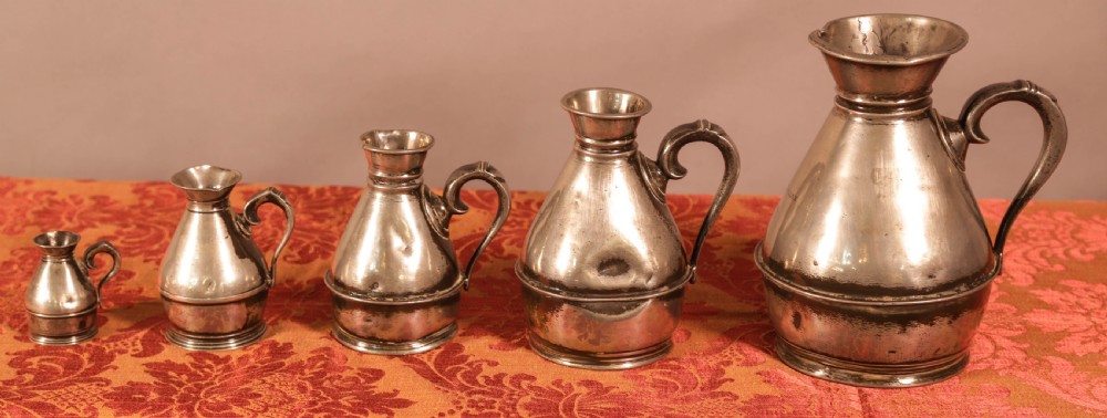 A graduated set of five antique pewter harvest jugs lined up on a patterned red surface. View VI