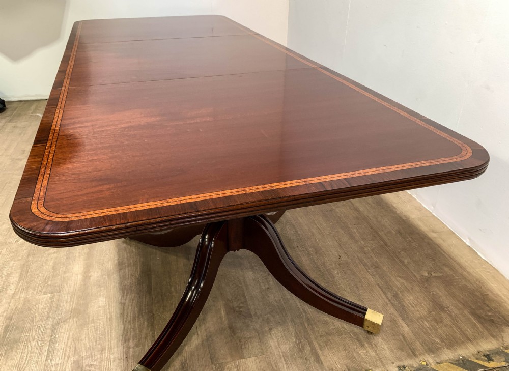 An angled view of a polished dark mahogany twin pedestal dining table featuring banded inlay and brass capped feet.