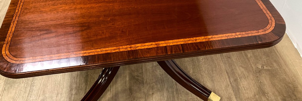 An angled view of a polished dark mahogany twin pedestal dining table featuring banded inlay and brass capped feet. View III