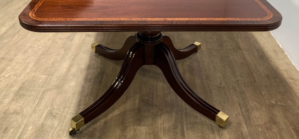 An angled view of a polished dark mahogany twin pedestal dining table featuring banded inlay and brass capped feet. View IV