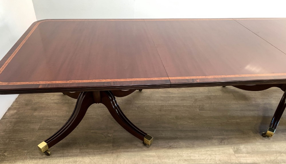 An angled view of a polished dark mahogany twin pedestal dining table featuring banded inlay and brass capped feet. View VI