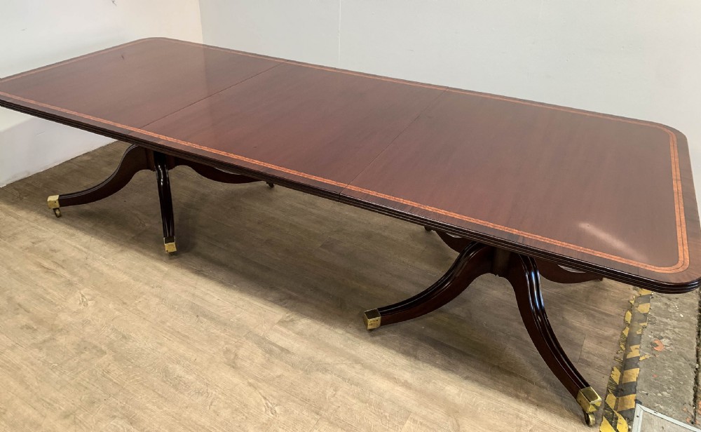 An angled view of a polished dark mahogany twin pedestal dining table featuring banded inlay and brass capped feet. View IX
