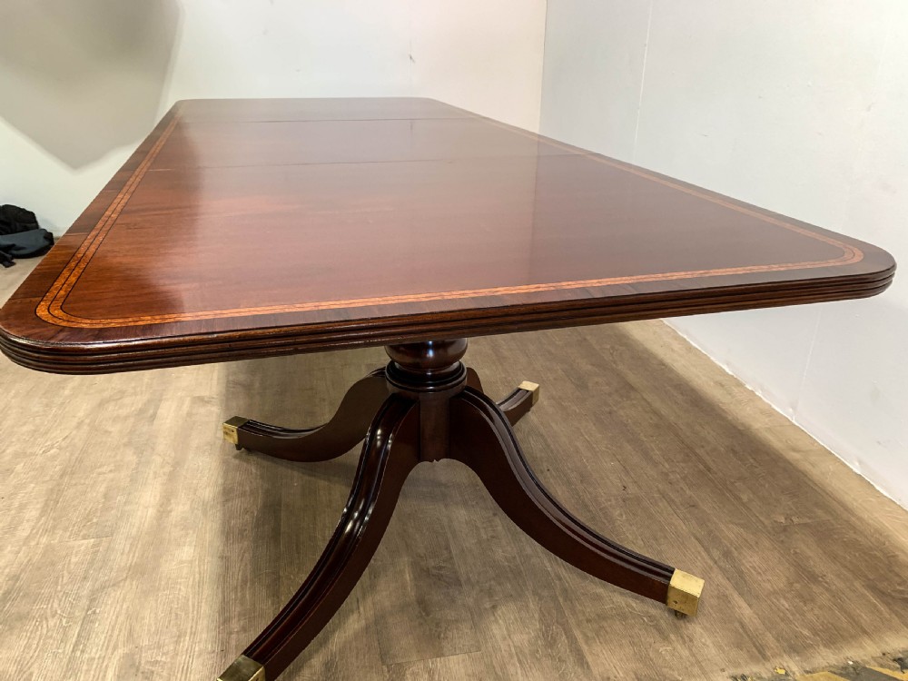 An angled view of a polished dark mahogany twin pedestal dining table featuring banded inlay and brass capped feet. View XI