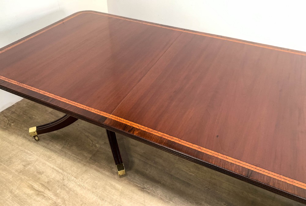An angled view of a polished dark mahogany twin pedestal dining table featuring banded inlay and brass capped feet. View XV