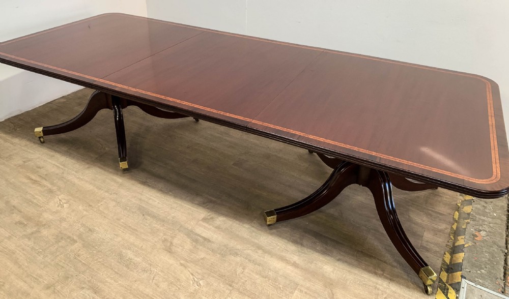 An angled view of a polished dark mahogany twin pedestal dining table featuring banded inlay and brass capped feet. View XVI