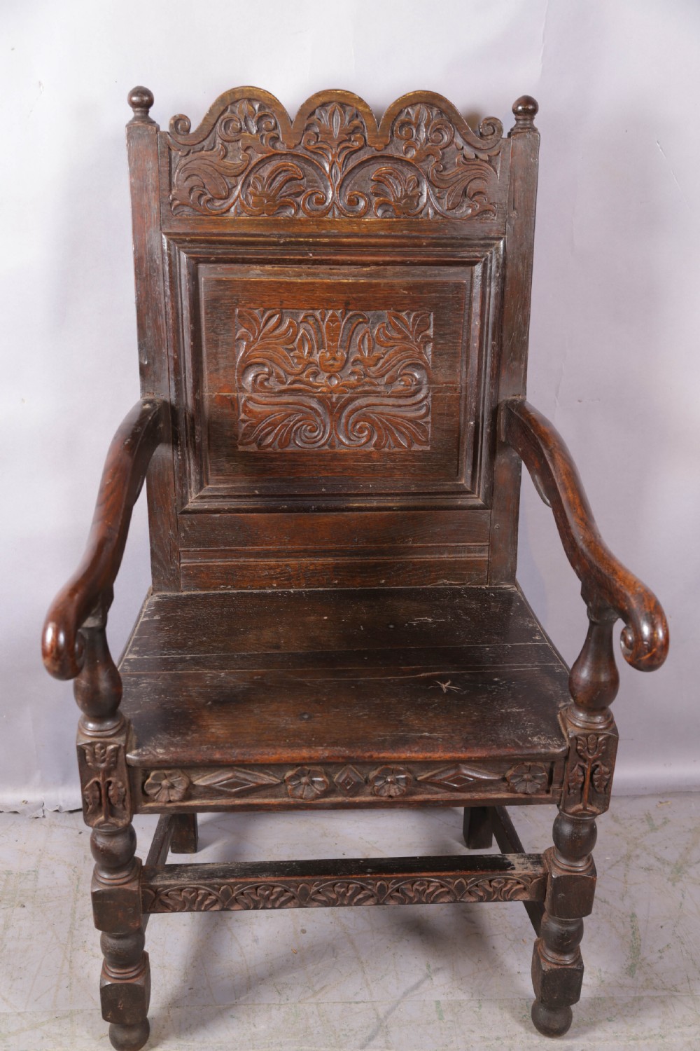 A dark oak wainscot chair featuring a carved panelled backrest, shaped scrolled arms, and turned front legs.
