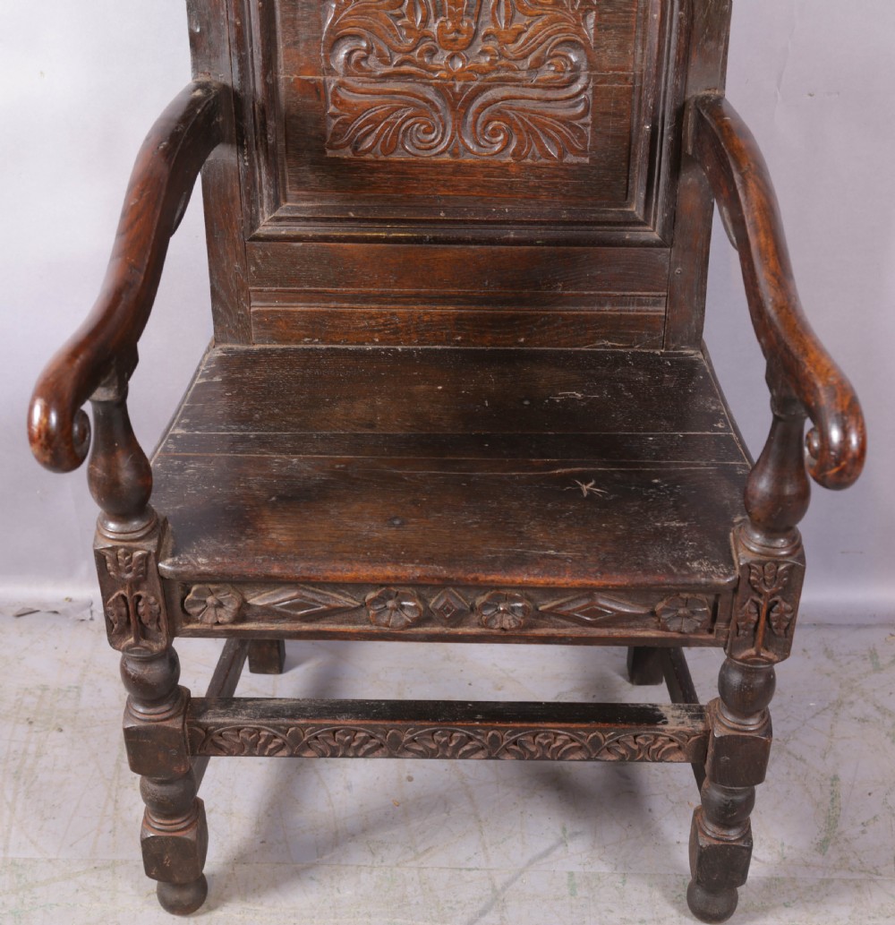 A dark oak wainscot chair featuring a carved panelled backrest, shaped scrolled arms, and turned front legs. View III
