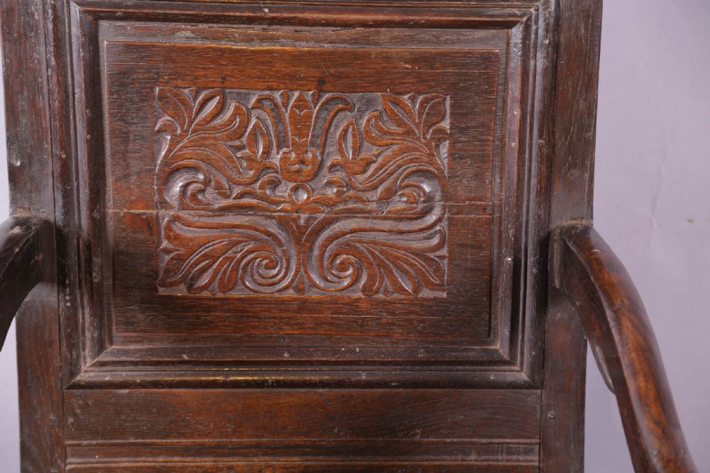 A dark oak wainscot chair featuring a carved panelled backrest, shaped scrolled arms, and turned front legs. View V