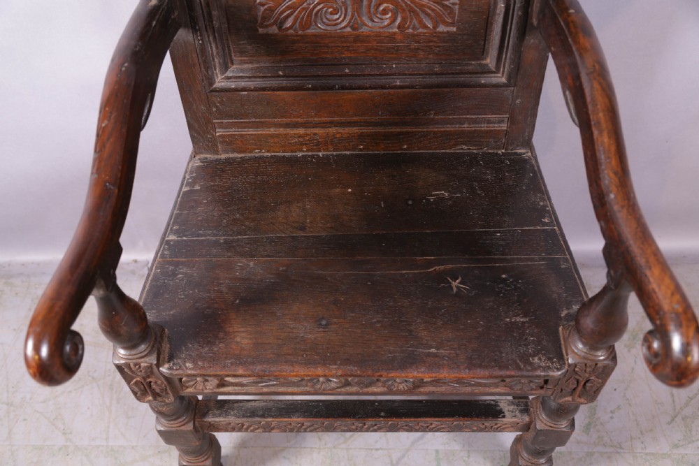 A dark oak wainscot chair featuring a carved panelled backrest, shaped scrolled arms, and turned front legs. View VI