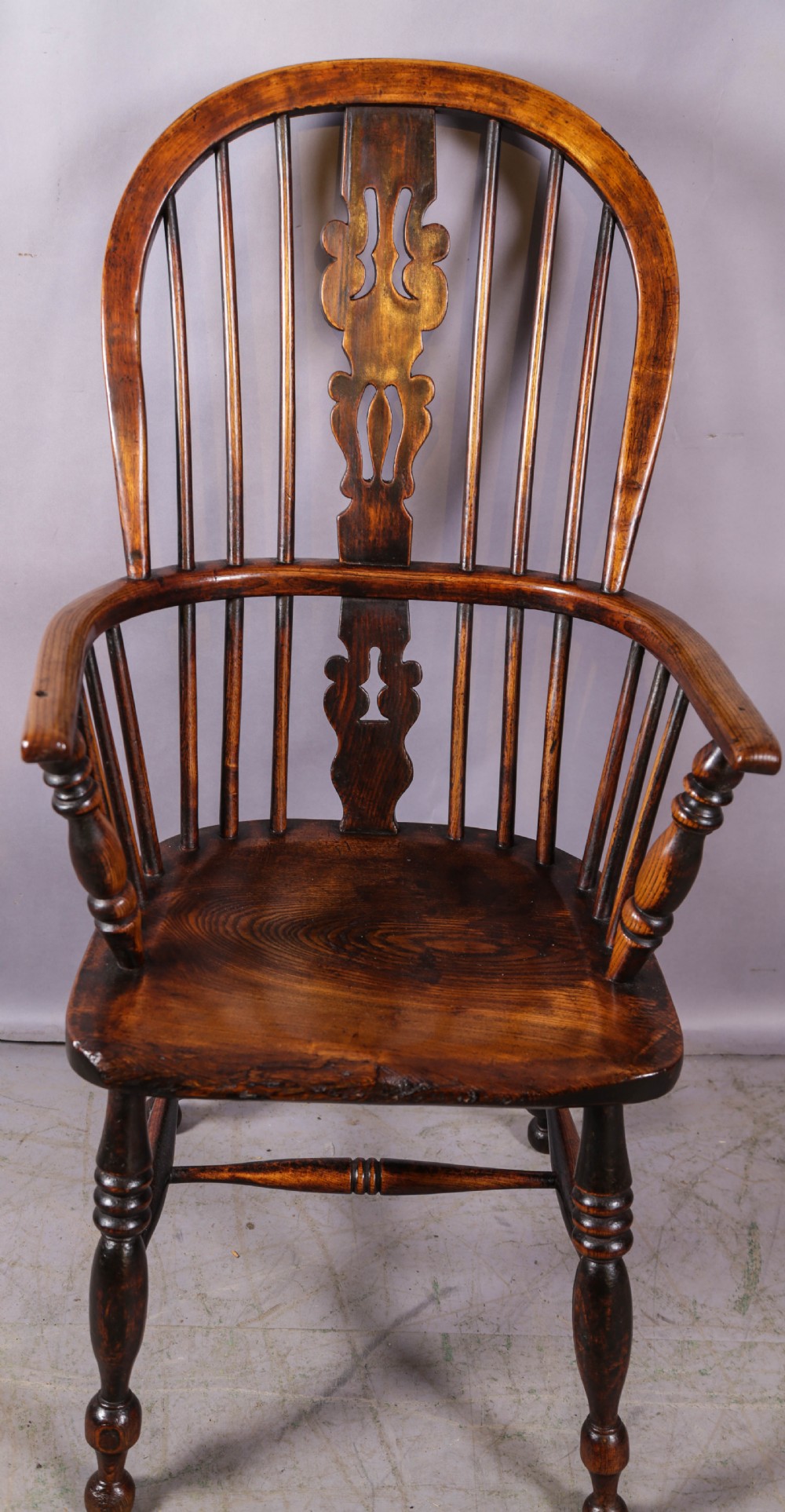 An antique high-back wooden Windsor armchair crafted from ash and elm with a pierced central splat, turned legs, and a shaped saddle seat.