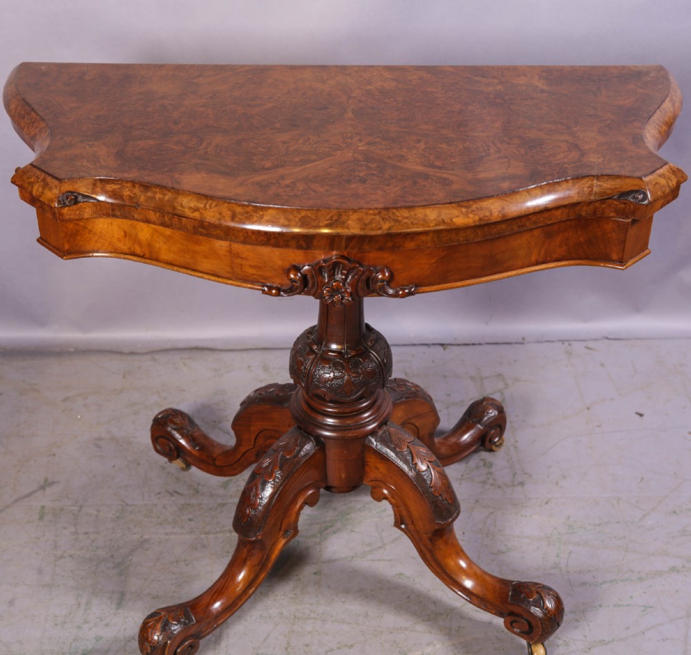A Victorian burr walnut fold-over card table featuring a shaped top, a carved pedestal stem, and carved quadripartite cabriole legs on brass castors.