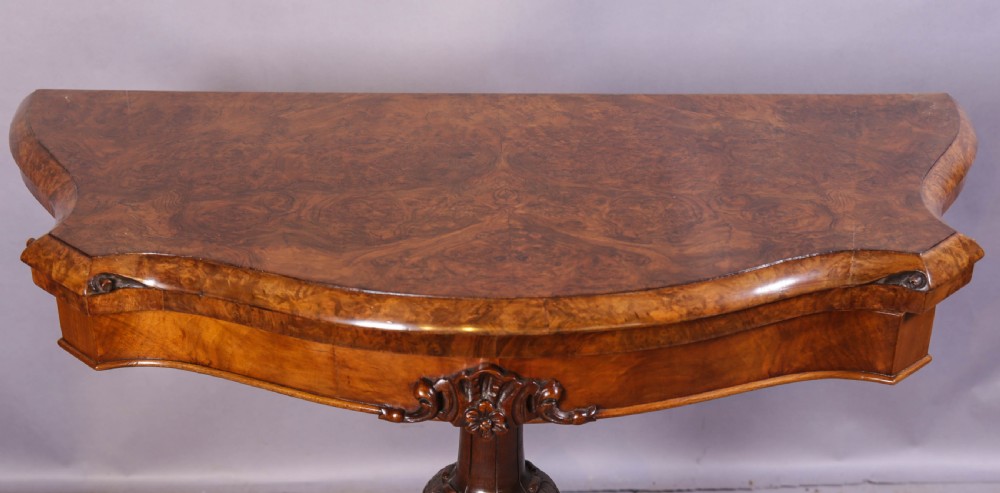 A Victorian burr walnut fold-over card table featuring a shaped top, a carved pedestal stem, and carved quadripartite cabriole legs on brass castors. View II