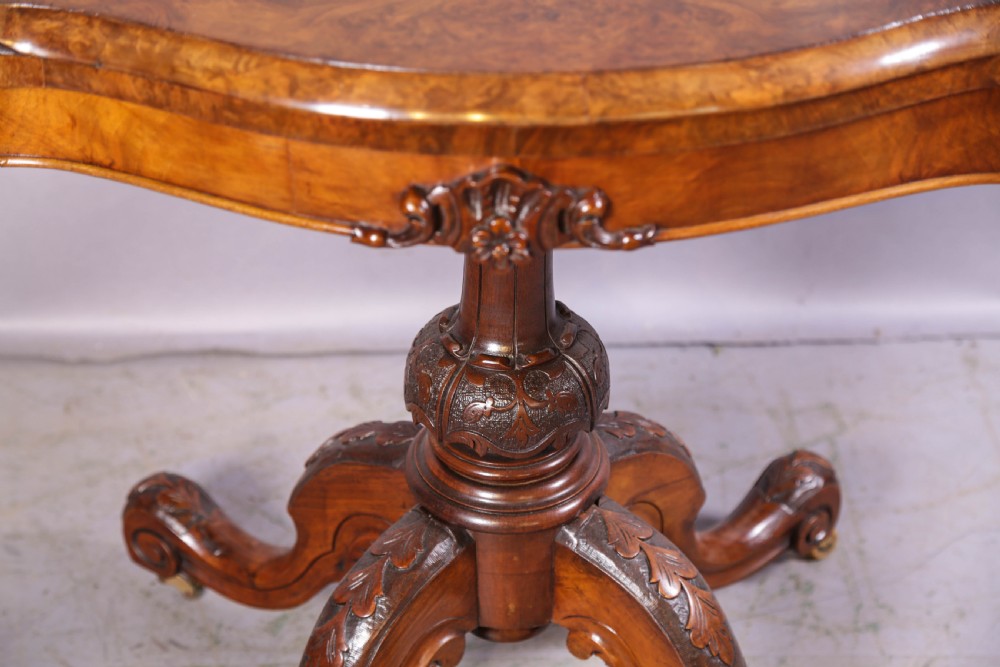 A Victorian burr walnut fold-over card table featuring a shaped top, a carved pedestal stem, and carved quadripartite cabriole legs on brass castors. View IV